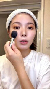 メイクブラシで頬にチークをのせて明るく笑う女性｜チーク｜メイク｜笑顔