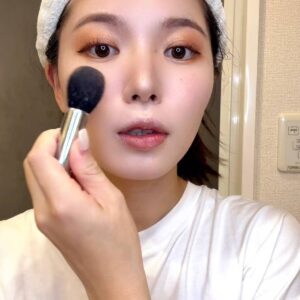 メイクブラシで頬にチークをのせて明るく笑う女性｜チーク｜メイク｜笑顔